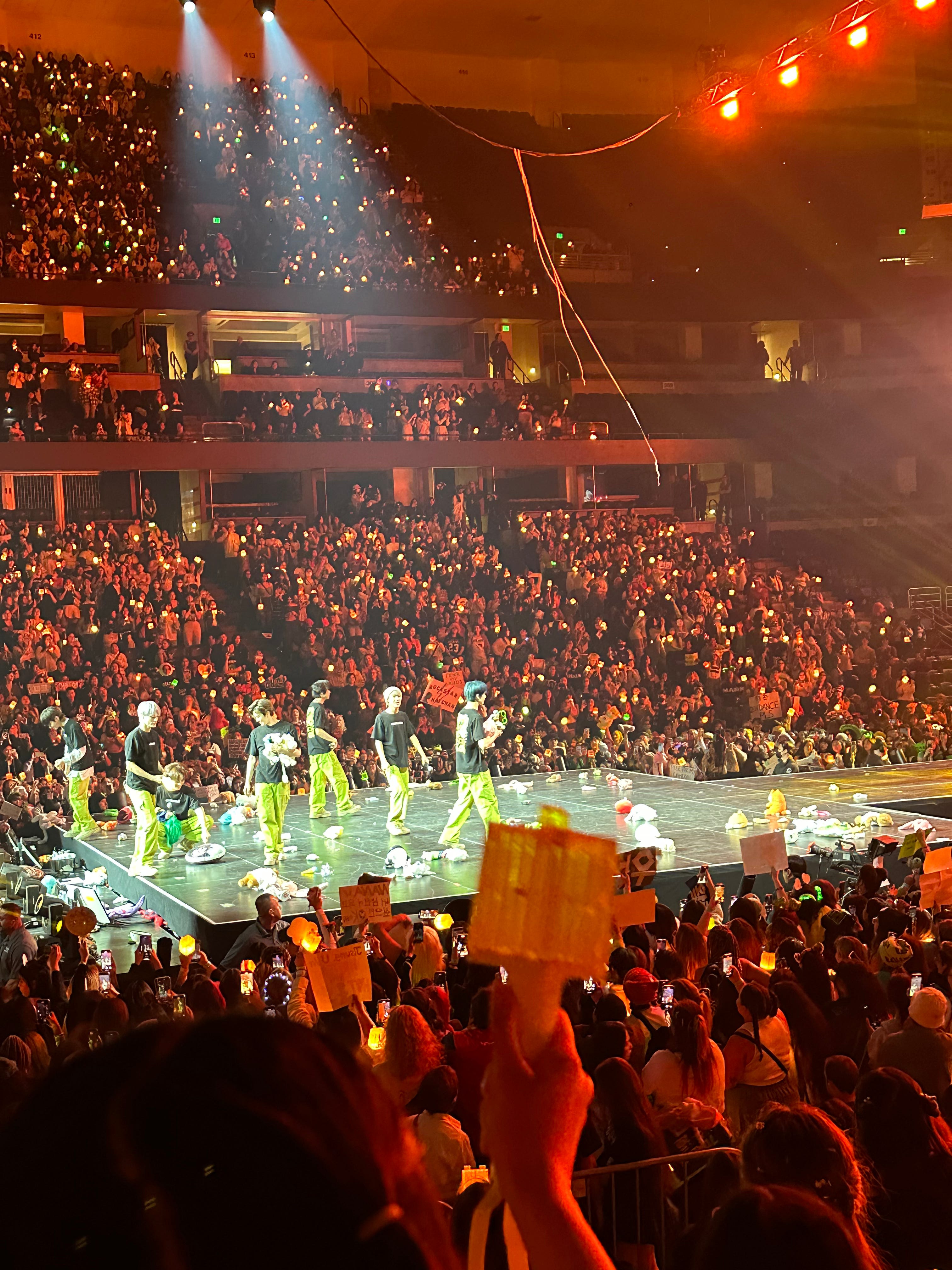 NCT Dream on stage, looking at stuffed animals fans threw on stage. Crowd is full, featuring fans in attendance at a concert, holding up green NCT lightsticks. One lightstick is in the forefront of the picture. 