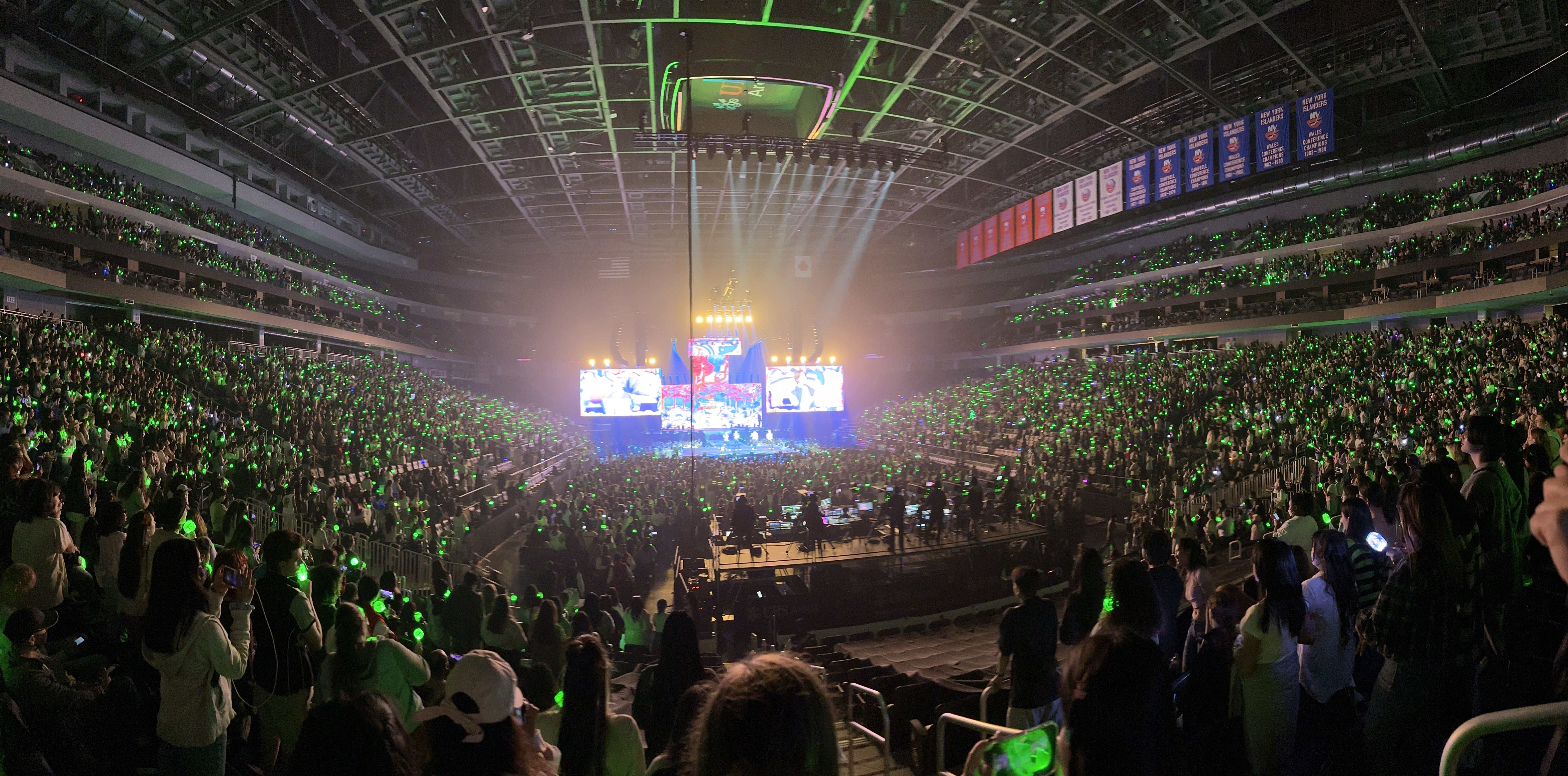 A panorama shot of Tomorrow X Together in concert, with crowd's lightsticks lit up in neon green