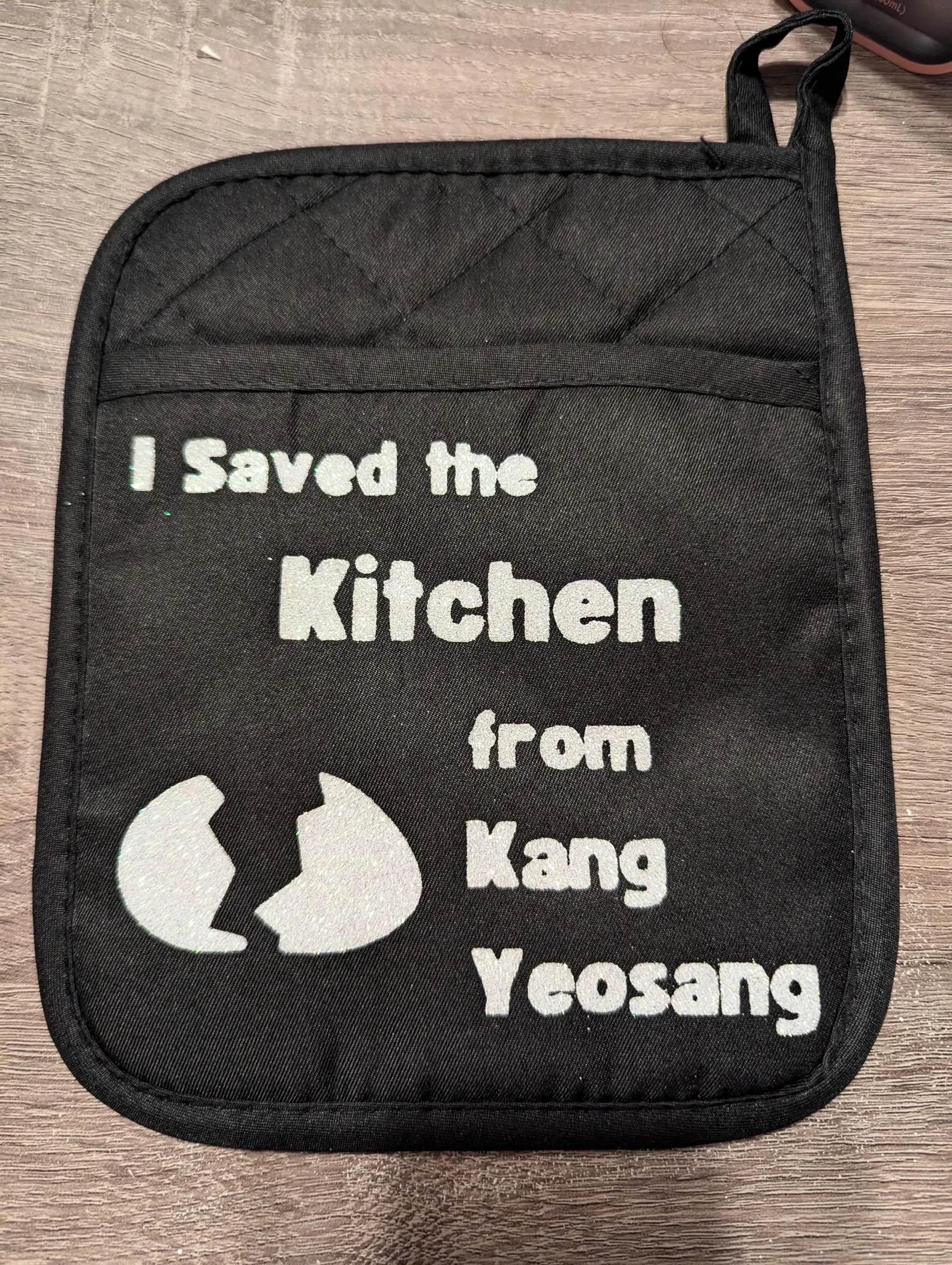 Black potholder with write graphics featuring a cracked egg with the phrase "I saved the kitchen from Kang Yeosang"