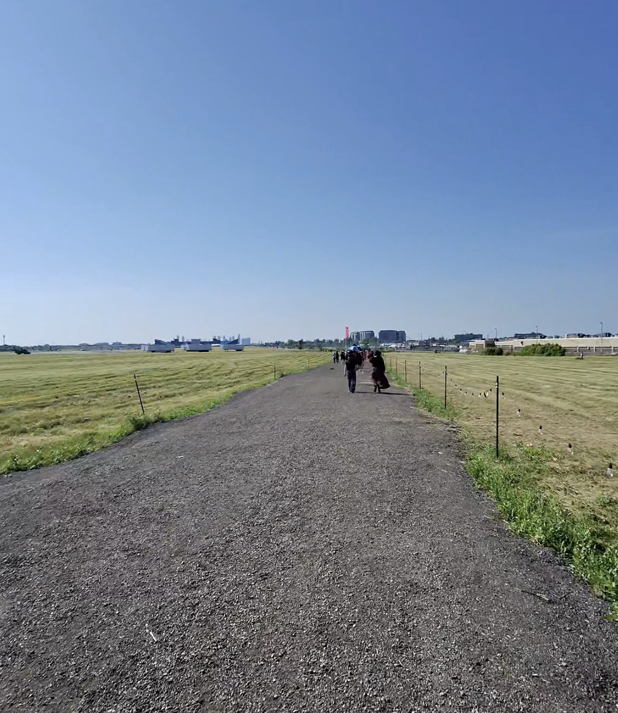 A picture of an asphalt road with people walking on it between fields of grass, surrounded by buildings very far off in the distance