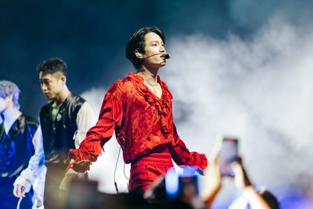 Kai in red on stage, with fog and two dancers, as a fan takes a video blurred in the crowd below.